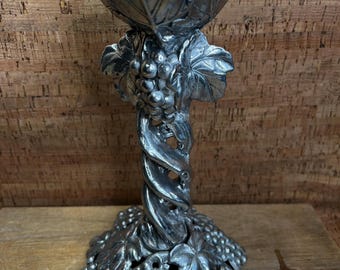 May include: A silver-colored decorative candle holder with a grape and leaf design. The base and top of the holder feature clusters of grapes and leaves. The stem is a twisted vine shape.