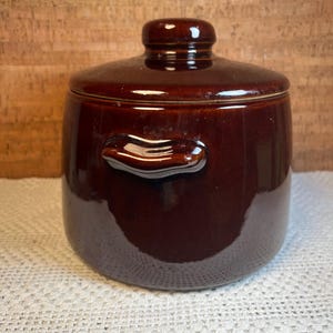 May include: A brown ceramic jar with a lid and two small handles. The jar has a glossy finish and is sitting on a white, crocheted surface. The background is a wooden panel.