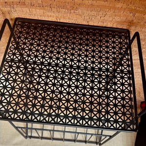 May include: A black metal side table with a geometric patterned top and a lower shelf. The table has a rectangular shape and is made of thin metal rods. The table is designed for indoor use.
