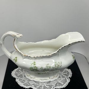 May include: White porcelain gravy boat with a handle and a scalloped rim, decorated with green floral sprigs and gold trim. The gravy boat sits on a white lace doily.