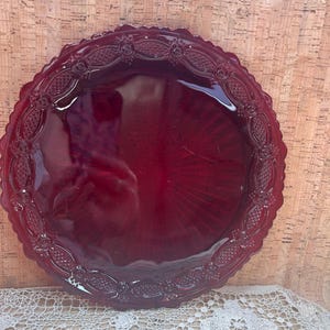 May include: A round, deep red glass plate featuring a decorative scalloped edge and a textured border pattern. The center of the plate is smooth and translucent, displaying a radial sunburst design that extends outward from the middle.