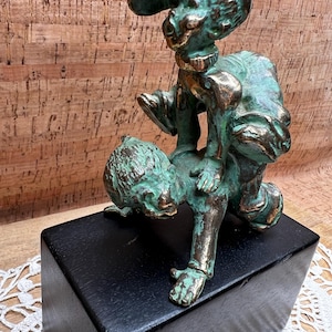 May include: A bronze sculpture depicting two figures in a playful pose. One figure, wearing a cap, is perched atop another. The sculpture has a verdigris patina, mounted on a black wooden base. The artwork is set against a textured, light brown background.