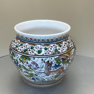 May include: A round, white ceramic pot with a wide opening. The pot is decorated with a colorful floral and animal design, featuring a deer. The rim has a detailed border. The pot is likely for decorative purposes.