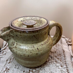 May include: A ceramic teapot with a speckled light green glaze and a brown rim. The teapot features a rounded body, a short spout, a curved handle, and a matching lid with a small central knob. The surface has a textured, earthy appearance.