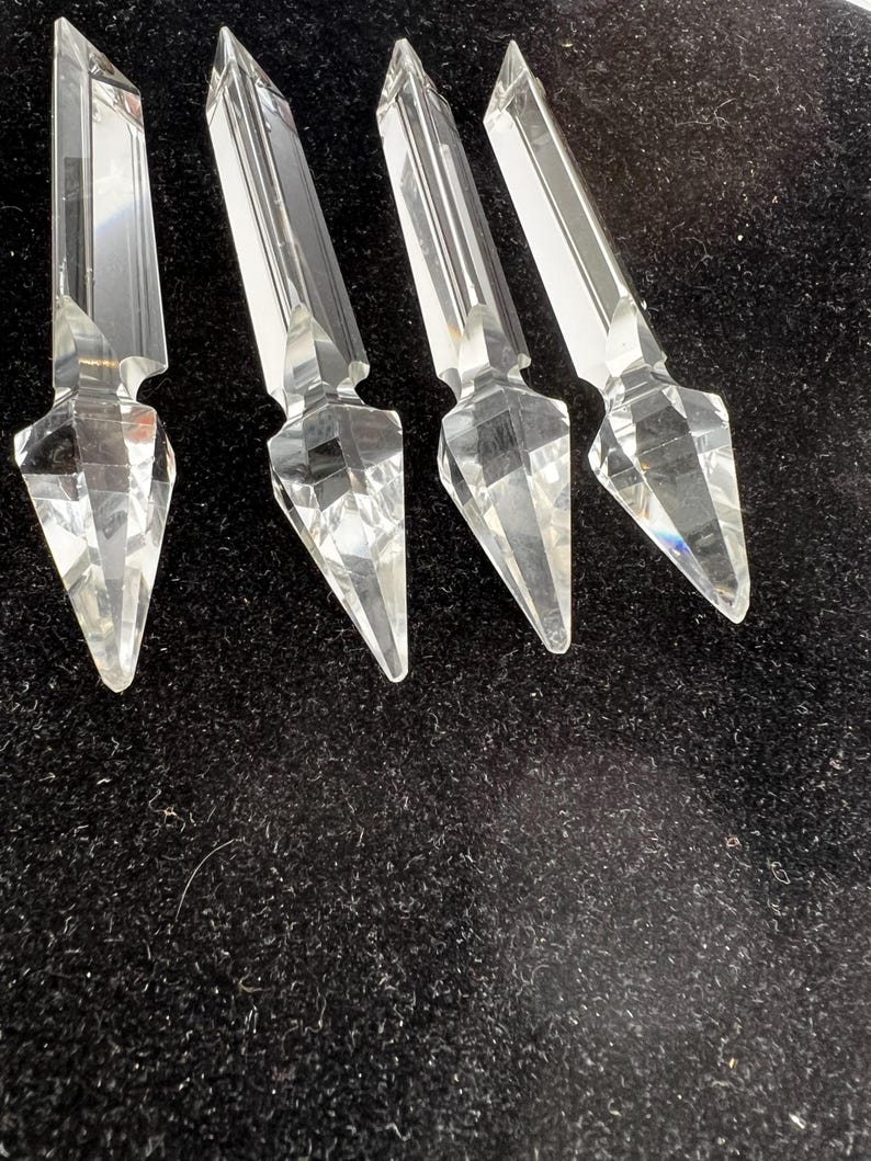 Puede incluir: Cuatro piezas decorativas de cristal transparente y facetado con un extremo puntiagudo y una parte superior rectangular. Los cristales est&aacute;n dispuestos sobre una superficie oscura y texturizada, mostrando sus cualidades reflectantes y dise&ntilde;o geom&eacute;trico.