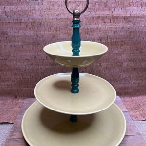 May include: A three-tiered serving stand with a brass handle and teal accents. The stand features three beige-colored plates, perfect for displaying desserts or appetizers. The stand is approximately 12 inches tall.