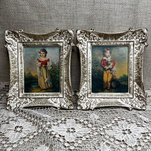 May include: Two antique framed portraits. Each features a child holding a small animal, set against a landscape. The frames are ornate, with a distressed silver finish. The artwork has a vintage aesthetic, with soft colors and detailed brushstrokes.