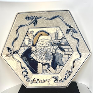 May include: Hexagonal ceramic plate with a Santa Claus design. The plate features a blue and white color scheme with a detailed illustration of Santa Claus holding a cookie and a stocking. The words "Cookies for Santa" are written on the plate.