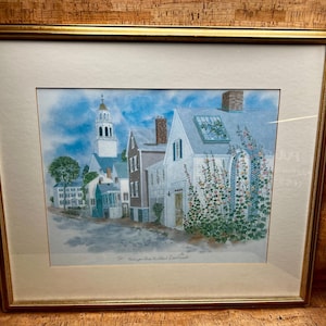 May include: Framed watercolor print of a New England town scene. The artwork depicts a white church with a steeple, colorful houses, and flowering plants. The print is set in a gold frame.