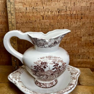 May include: White and brown ceramic pitcher and basin set. The pitcher has a curved handle and a spout with a scalloped edge. The set features a brown landscape design. The basin has a scalloped edge and a floral design on the base.
