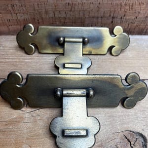 May include: Two antique brass drawer pulls with decorative, curved edges and a central hinged handle. The pulls are rectangular with a slightly aged appearance, set against a wooden background.