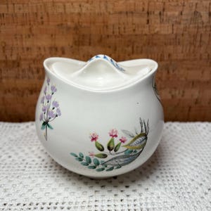 May include: White ceramic sugar bowl with a lid. The bowl has a floral design with pink, purple, and green flowers and leaves.