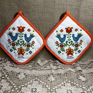May include: Two white, square Corning pot holders with an orange polka dot trim. Each pot holder features a floral and bird design in blue, red, and green. The text "©1975 CORNING" is printed on each pot holder.