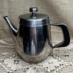 May include: A stainless steel teapot with a lid and a curved handle. The teapot has a spout and a small knob on top of the lid. The teapot is sitting on a lace doily.