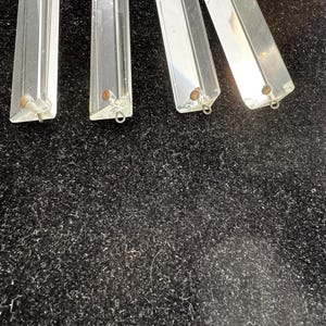 Puede incluir: Prismas de ara&ntilde;a de cristal transparente y rectangular con bordes facetados. Cada prisma tiene un peque&ntilde;o lazo de metal en la parte inferior para la fijaci&oacute;n. Los prismas se muestran sobre un fondo oscuro y texturizado.