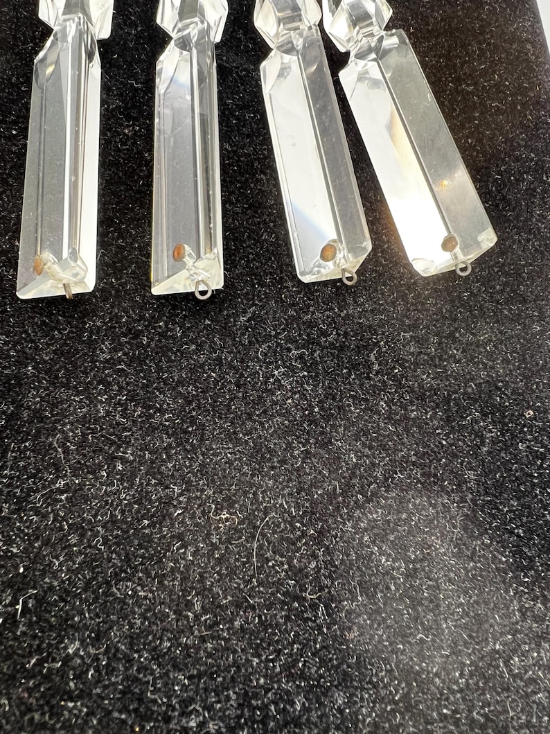 Puede incluir: Prismas de ara&ntilde;a de cristal transparente y rectangular con bordes facetados. Cada prisma tiene un peque&ntilde;o lazo de metal en la parte inferior para la fijaci&oacute;n. Los prismas se muestran sobre un fondo oscuro y texturizado.