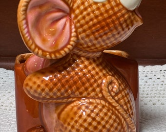 May include: A brown ceramic mouse planter with a pink ear and a white nose. The mouse is holding a brown pot with a checkered pattern.