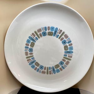 May include: A white ceramic plate with a circular design of geometric shapes in blue, brown, and teal. The plate has a wide rim and a smooth, glossy finish. This vintage plate is perfect for serving food.