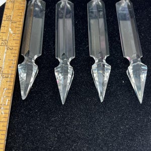 May include: Four clear, faceted crystal prisms with pointed ends, arranged on a black surface. Each prism is approximately 3 inches long. The prisms have a rectangular body and a decorative, geometric design.