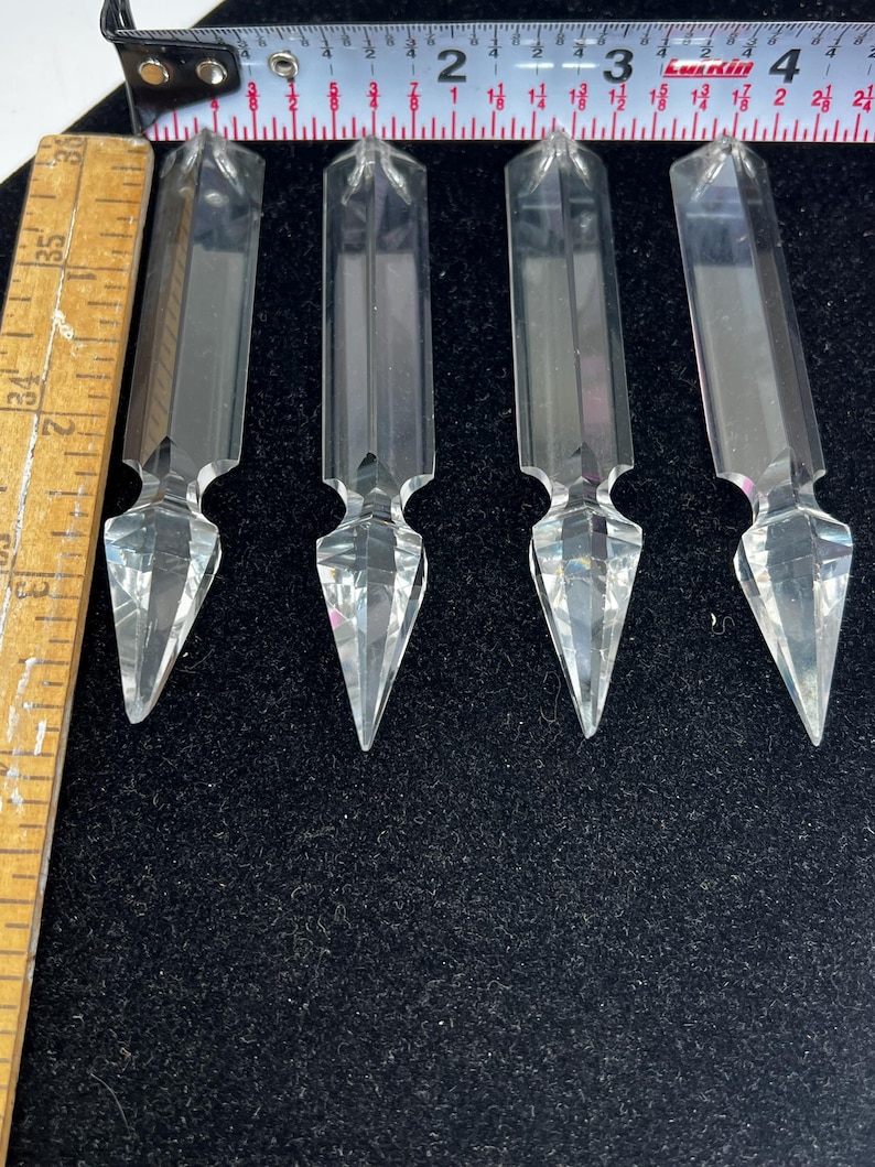 Puede incluir: Cuatro prismas de cristal transparente y facetado con extremos puntiagudos, dispuestos sobre una superficie negra. Cada prisma mide aproximadamente 7,6 cm de largo. Los prismas tienen un cuerpo rectangular y un dise&ntilde;o geom&eacute;trico decorativo.