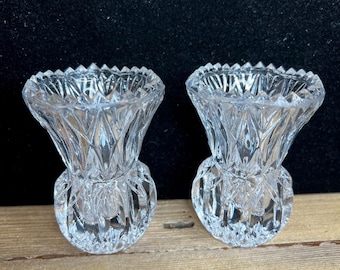 May include: Two clear, cut-glass thistle-shaped candle holders. Each has a round base, a textured body, and a flared top with a jagged edge. The candle holders are displayed on a wooden surface against a dark background.