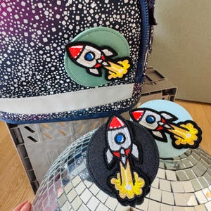 Könnte beinhalten: Drei bunte Raketen-Patches mit gestickten Details. Die Patches befinden sich auf einem blau-weiß gemusterten Hintergrund, einem schwarzen Hintergrund und einem hellblauen Hintergrund.