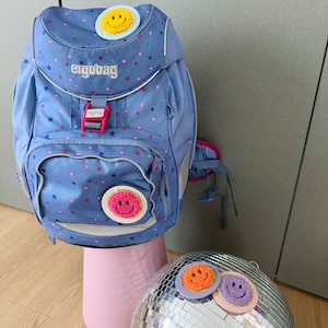 Könnte beinhalten: Ein blauer und rosa Rucksack mit einem weißen Sternenmuster und dem Wort "ergobag" auf der Vorderseite. Der Rucksack hat drei gelbe, orange und rosa Smiley-Patches.