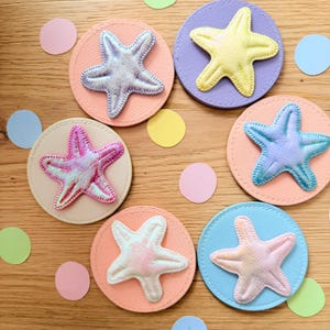 Könnte beinhalten: Sechs dekorative Untersetzer mit Seestern-Designs. Jeder Untersetzer hat einen genähten Seestern in Pastellfarben, darunter Rosa, Blau und Gelb, auf einem kreisförmigen Hintergrund. Die Untersetzer sind auf einer Holzoberfläche mit Konfetti angeordnet.