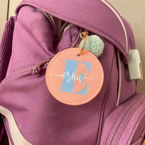 Könnte beinhalten: Ein runder rosa Schlüsselanhänger mit dem Buchstaben E in Blau und dem Wort "erha" in weißer Schrift. Der Schlüsselanhänger ist an einem lila Rucksack befestigt.