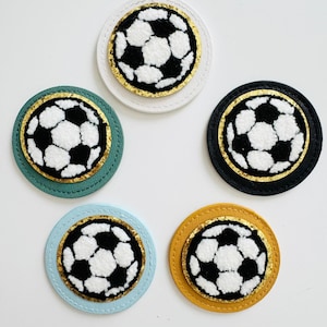 Könnte beinhalten: Fünf runde Filzpatches mit goldener Umrandung und einem weißen und schwarzen Fußballdesign. Die Patches befinden sich auf einem weißen Hintergrund. Die Patches sind in verschiedenen Farben: weiß, grün, schwarz, blau und gelb.