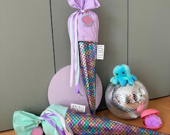 Könnte beinhalten: Zwei Schultüten im Meerjungfrauen-Design, eine aufrecht und eine liegend, mit schillernden, schuppenartigen Schwänzen. Die aufrechte Tüte ist lavendelfarben mit dem Namen "LOTTI" auf einem Etikett. Die andere ist mintgrün mit dem Namen "ANNI". Eine Discokugel und ein Plüsch-Oktopus sind ebenfalls vorhanden.