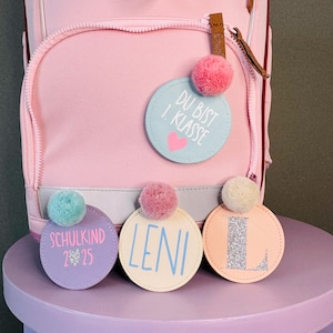 Könnte beinhalten: Vier bunte runde Stoffanhänger mit Bommeln. Die Anhänger sagen "Du bist 1. Klasse", "Schulkind 2/25", "Leni" und "L".