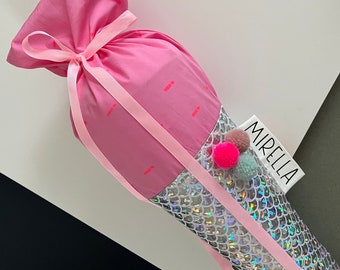 May include: A pink fabric cone with a white tag that says "Mirella" and pink and green pom-poms. The cone is decorated with a silver mermaid scale pattern.