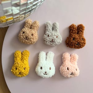 Könnte beinhalten: Sechs Hasenkopf-Ornamente aus Plüsch in verschiedenen Farben. Die Hasen sind in Braun-, Grau-, Gelb-, Weiß- und Rosatönen gehalten. Jeder Hase hat gestickte Gesichtszüge und ein flauschiges Aussehen.