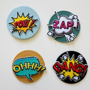 Könnte beinhalten: Vier runde gestickte Patches mit verschiedenen Comic-Soundeffekten. Die Patches befinden sich auf einem weißen Hintergrund. Die Patches sind: "POW!" auf einem hellblauen Hintergrund, "ZAP!" auf einem grünen Hintergrund, "OHHH!" auf einem gelben Hintergrund und "BANG!" auf einem schwarzen Hintergrund.