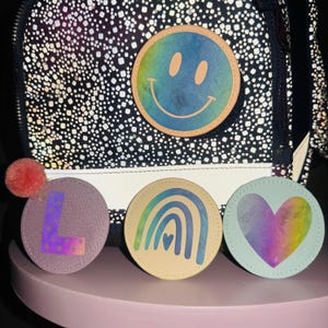 Könnte beinhalten: Vier bunte Stoffpatches mit verschiedenen Designs. Die Patches sind rund und zeigen einen Regenbogen, ein Herz, ein Smiley und einen Buchstaben L. Die Patches befinden sich alle auf einem schwarz-weißen Hintergrund.