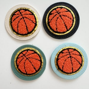 Könnte beinhalten: Vier gestickte Filzpatches mit einem Basketball-Motiv. Die Patches sind in verschiedenen Farben: weiß, schwarz, hellblau und grün. Jeder Patch hat einen goldenen Umriss.