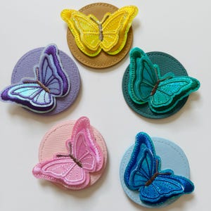 Könnte beinhalten: Fünf runde Filzscheiben mit gestickten Schmetterlingen in verschiedenen Farben. Die Schmetterlinge sind gelb, lila, grün, rosa und blau.