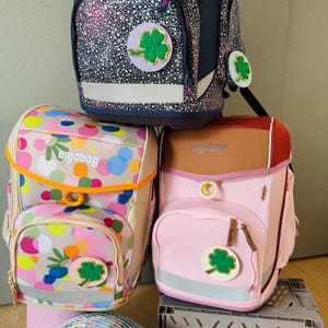 Könnte beinhalten: Drei farbenfrohe Rucksäcke mit dem Markennamen "ergobag" darauf gedruckt. Die Rucksäcke sind übereinander gestapelt. Jeder Rucksack hat einen grünen Kleeblatt-Aufnäher.