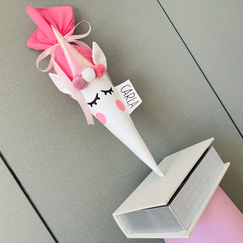 K&ouml;nnte beinhalten: Eine wei&szlig;e Geschenk-T&uuml;te im Einhorn-Design mit rosa Akzenten, einem glitzernden Horn und dem Namen "CARLA" auf einem Etikett. Der Kegel wird mit einer kleinen, cremefarbenen, buchf&ouml;rmigen Schachtel und einer rosa Stoffverpackung kombiniert.