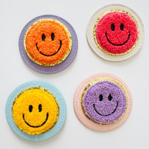 Könnte beinhalten: Vier gestickte Smiley-Patches mit verschiedenen Hintergrundfarben. Die Patches sind orange, rot, lila und gelb. Jeder Patch hat einen goldenen Glitzerrand.