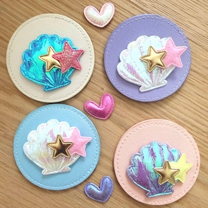 Könnte beinhalten: Vier runde Stoffaufnäher mit Muschel- und Sterndesigns. Jeder Aufnäher hat einen Hintergrund in einer anderen Farbe: Beige, Lavendel, Hellblau und Rosa. Die Muscheln haben schillernde Akzente, und die Sterne sind gold- und rosafarben. Kleine herzförmige Verzierungen sind verstreut.