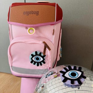 Könnte beinhalten: Rosa Rucksack mit einer braunen Lederklappe und dem Wort "ergobag" darauf. Der Rucksack hat einen blauen und weißen Augen-Patch auf der Vorderseite. Eine silberne Discokugel mit einem blauen und weißen Augen-Patch darauf befindet sich im Vordergrund.