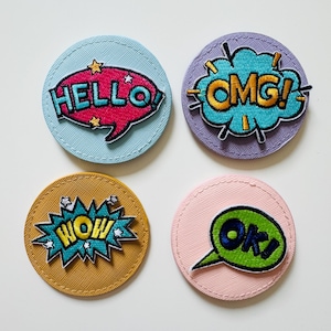 Könnte beinhalten: Vier runde, bunte Filzpatches mit gestickten Sprechblasen. Die Patches zeigen die Wörter "HELLO", "OMG", "WOW" und "OK" im Comic-Stil.