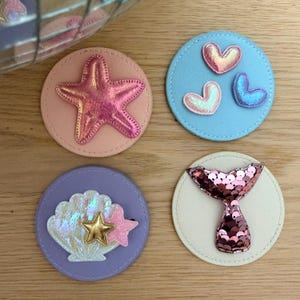 Könnte beinhalten: Vier runde, dekorative Aufnäher mit Meerjungfrauen- und maritimen Themen. Ein Aufnäher zeigt einen rosa Seestern auf pfirsichfarbenem Hintergrund. Ein anderer hat drei schillernde Herzen auf blauem Hintergrund. Ein dritter Aufnäher zeigt eine Muschel mit einem goldenen Stern und einem rosa Stern. Der letzte hat eine rosa Pailletten-Meerjungfrauenflosse.