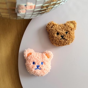 Könnte beinhalten: Zwei Aufnäher mit Bärenkopf aus Plüsch. Einer ist hellbraun, der andere rosa. Beide haben gestickte Merkmale, darunter schwarze Augen und eine schwarze Nase. Der rosa Bär hat orangefarbene Ohren und blaue Akzente. Der hellbraune Bär hat braune Akzente.