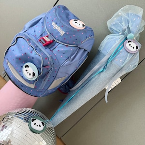 Könnte beinhalten: Ein blauer Rucksack mit Sternenmuster und dem Aufdruck "ergobag". Der Rucksack hat einen Pandabären-Aufnäher auf der Vorderseite. Außerdem gibt es einen blauen und weißen Kegel mit einem Pandabären-Aufnäher.