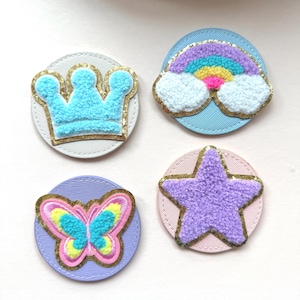 Könnte beinhalten: Vier farbenfrohe, gestickte Aufnäher. Designs umfassen eine hellblaue Krone, einen Regenbogen mit weißen Wolken, einen Schmetterling mit rosa, gelben und blauen Flügeln und einen lila Stern. Jeder Aufnäher hat einen kreisförmigen Hintergrund mit goldenem Rand.