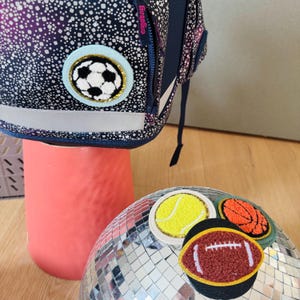 Könnte beinhalten: Vier gestickte Bügelbilder mit verschiedenen Sportbällen: ein Fußball, ein Tennisball, ein Basketball und ein American Football. Die Patches befinden sich auf einer silbernen Discokugel.