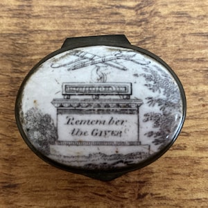 May include: A small, oval, silver-toned trinket box with a white porcelain lid. The lid features a black and white illustration of a monument with the text "Remember the Giver".